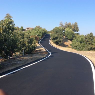 A freshly paved winding black road with white edge lines, bordered by trees on a sunny day.