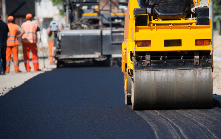 A yellow steamroller compacts fresh black asphalt on a road construction site, with workers standing in the background.