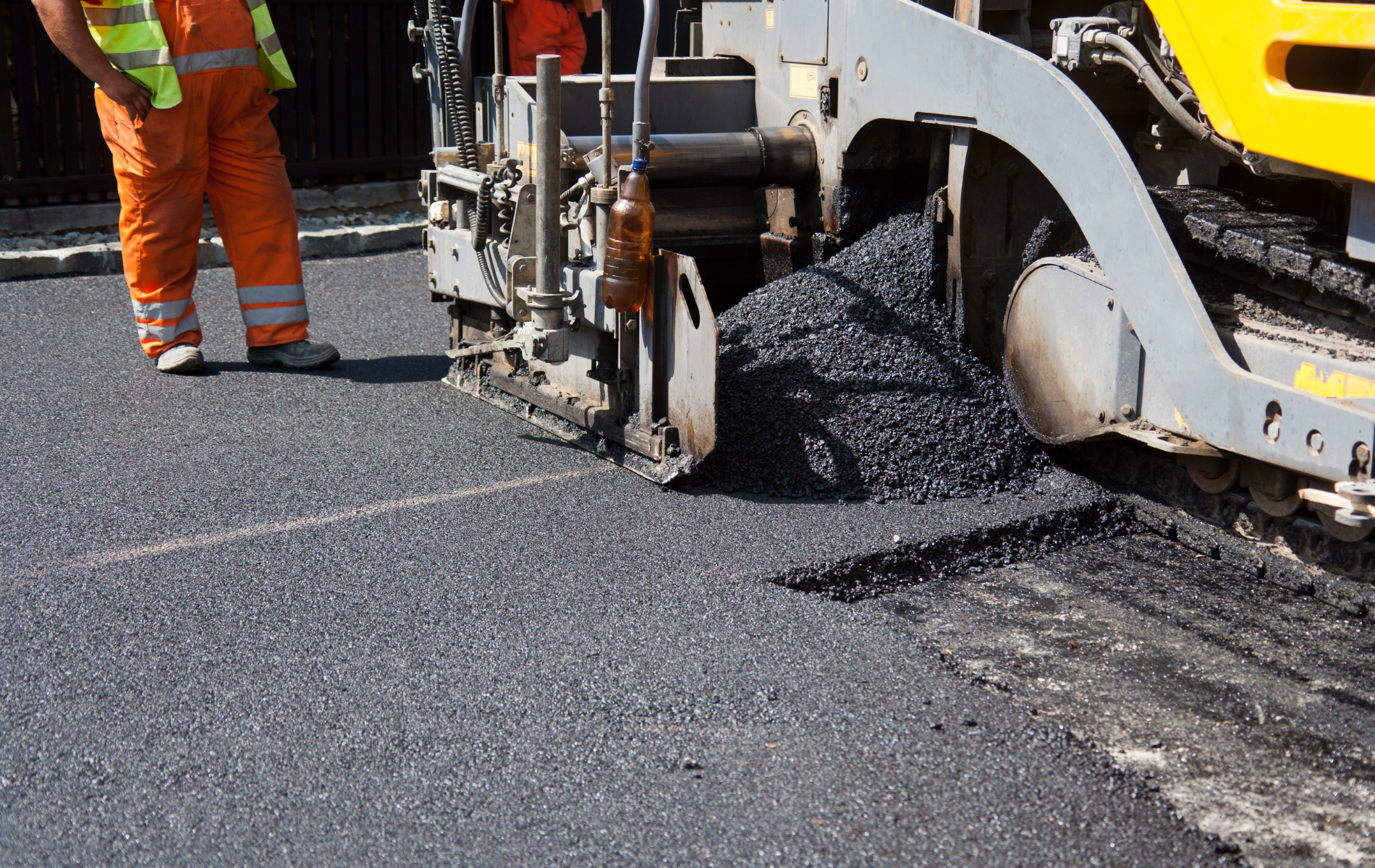A construction worker in orange high-visibility gear stands beside a road paver laying fresh black asphalt.