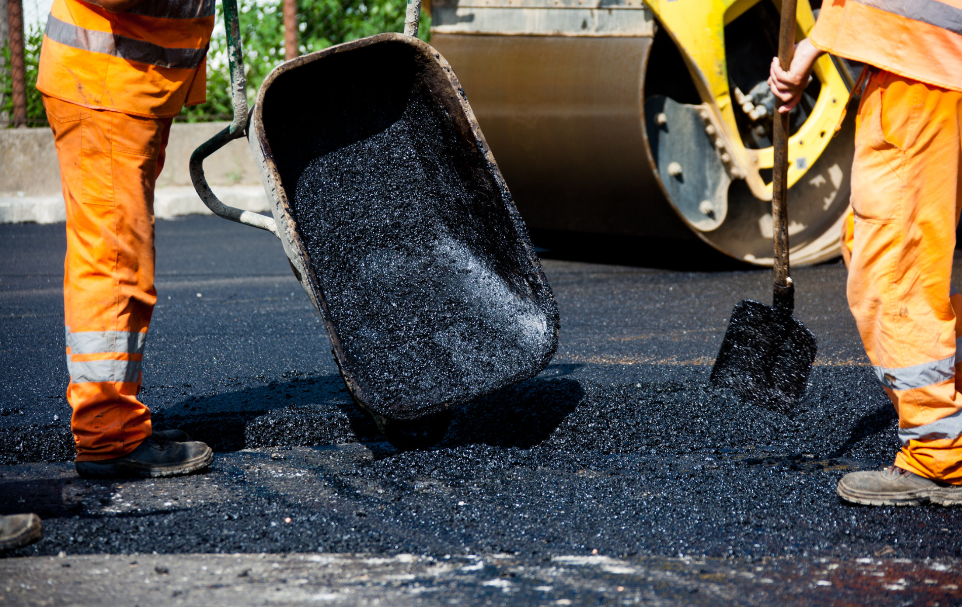 Two workers in orange high-visibility clothing dump asphalt from a wheelbarrow onto a road surface near a steamroller.