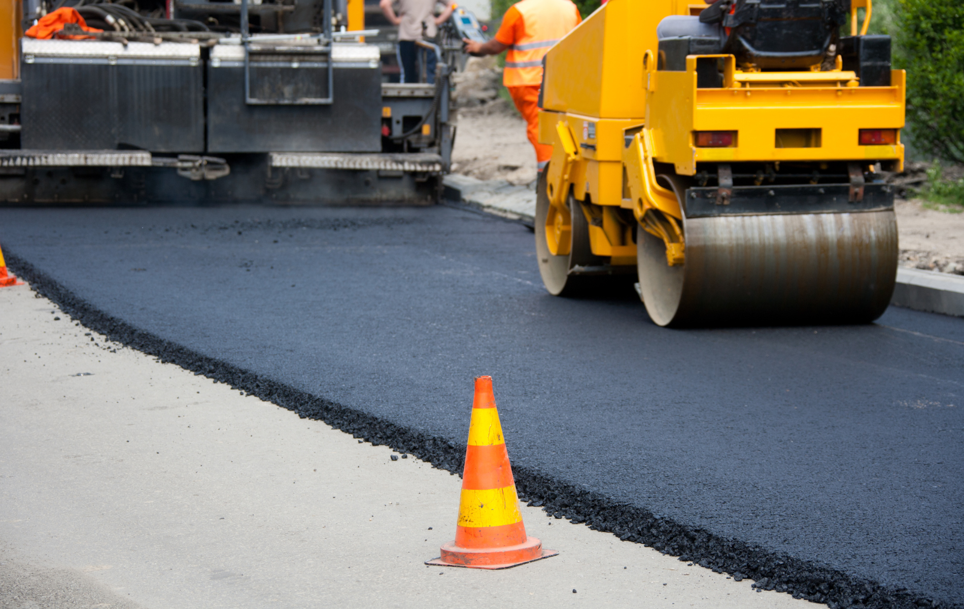 Road crew operating a steamroller to pave a fresh asphalt road, with an orange traffic cone in the foreground.