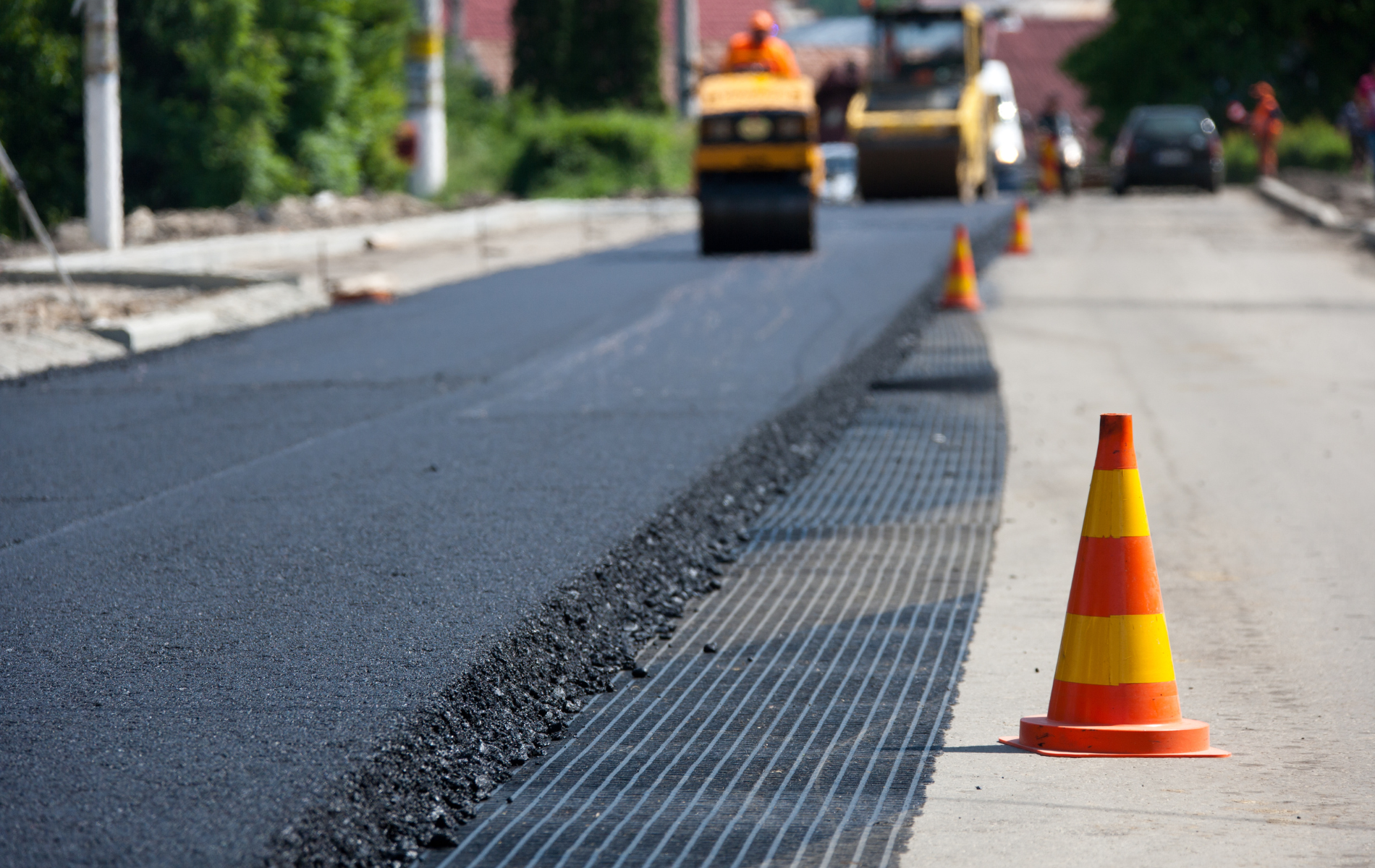 A construction site showing fresh black asphalt being paved next to a grid reinforcement layer with orange traffic cones.