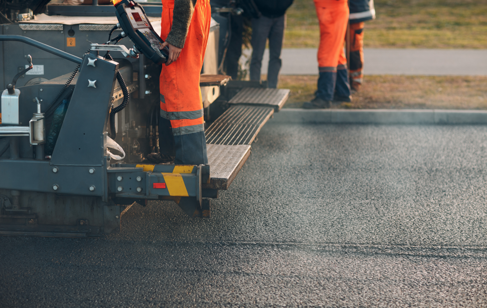 Road construction workers in orange high-visibility clothing operate an asphalt paving machine on a road surface.