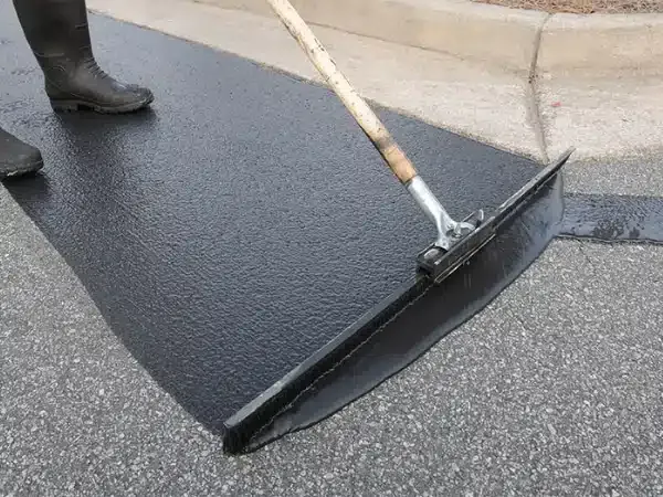 A worker uses a squeegee to apply black asphalt sealer to a paved driveway next to a concrete curb.
