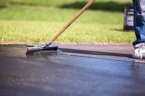 A person uses a squeegee to spread black asphalt sealant across a driveway.
