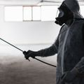 Personne en combinaison de protection pulvérisant du désinfectant. Intérieur, murs blancs, faiblement éclairé, respirateur sombre.