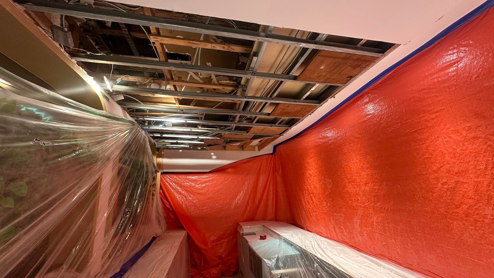Vue intérieure du plafond endommagé avec poutres apparentes, entouré de bâches en plastique rouges et transparentes.