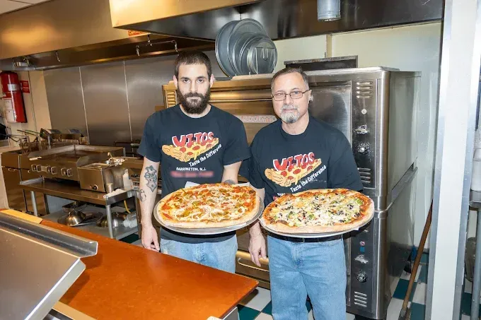 Two men in a pizza kitchen hold pizzas. One has a beard. Stainless steel oven in the background.
