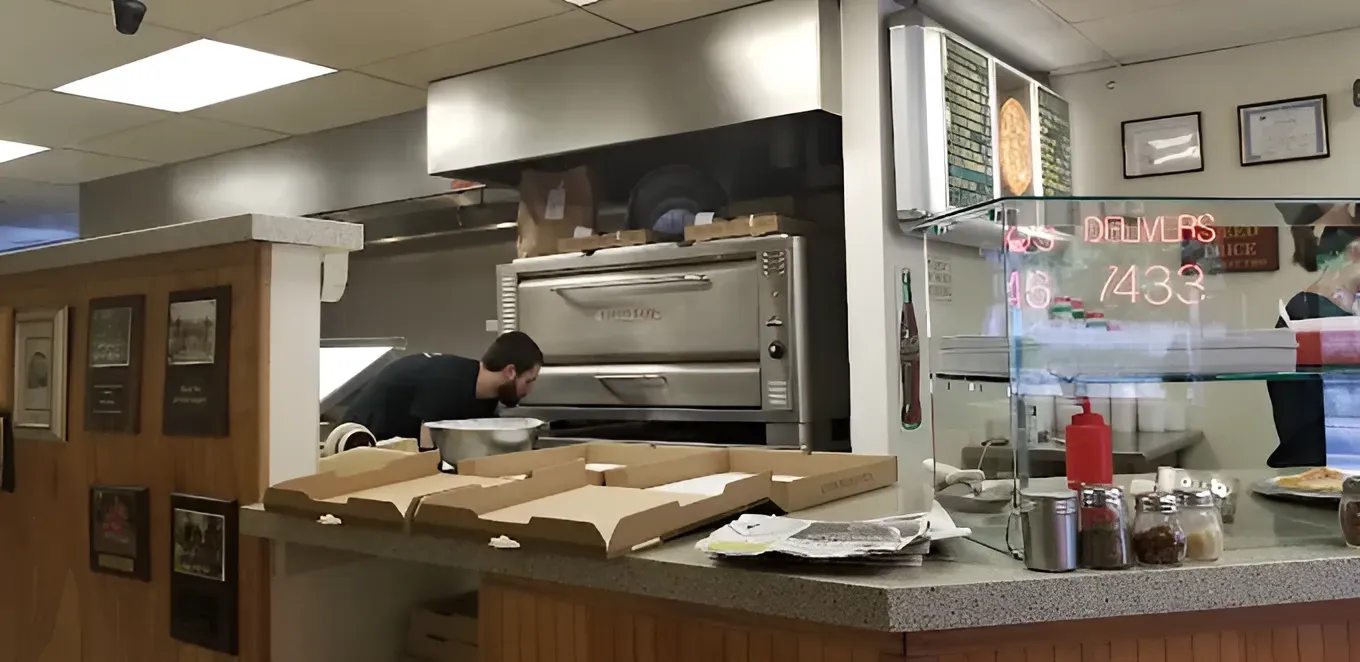 A man takes pizza from an oven in a restaurant. Pizza boxes are on the counter.