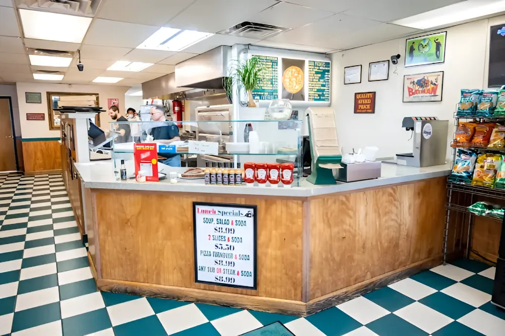 Counter and kitchen of a pizza restaurant with checkered floor, menu, and chip rack.