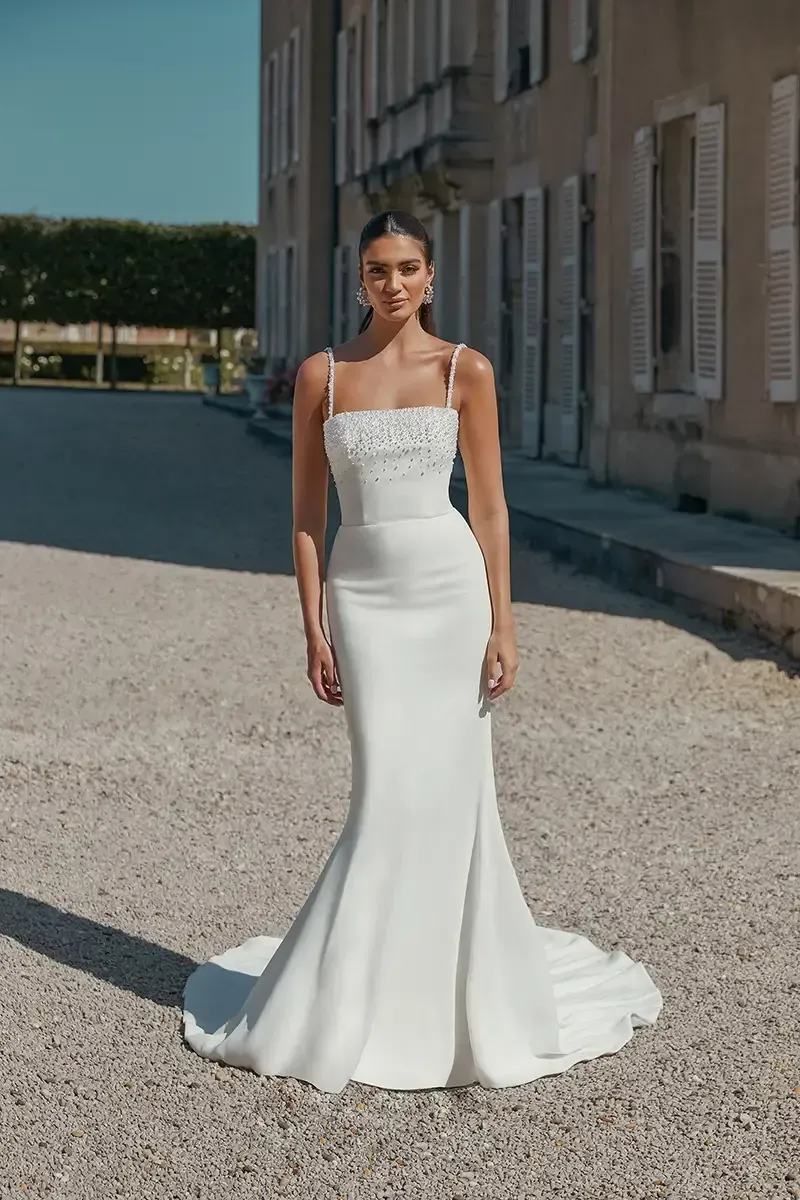 Woman in white wedding dress with square neckline and train, standing outside a building.