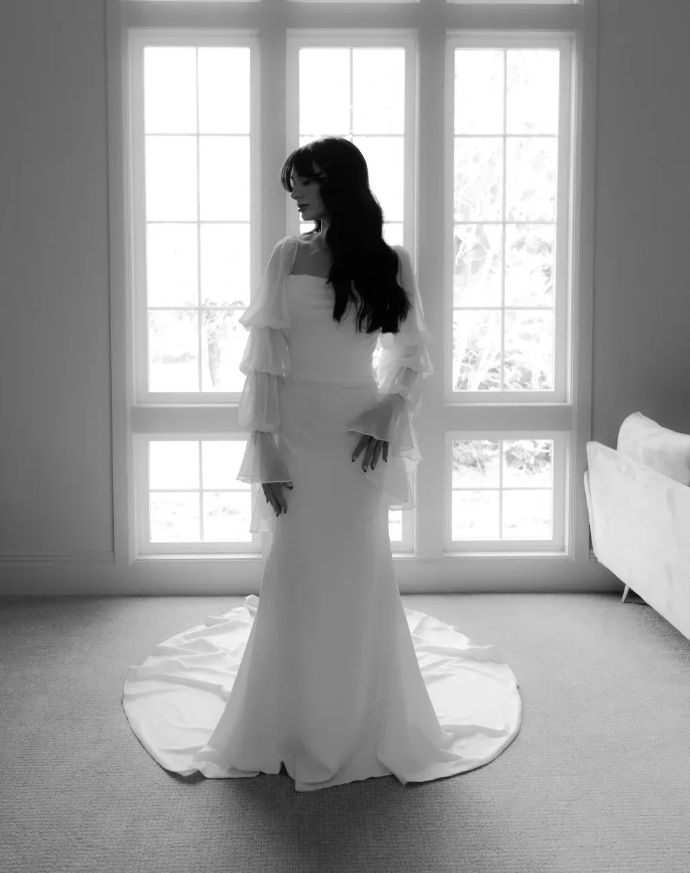 Woman in white wedding dress stands before window.