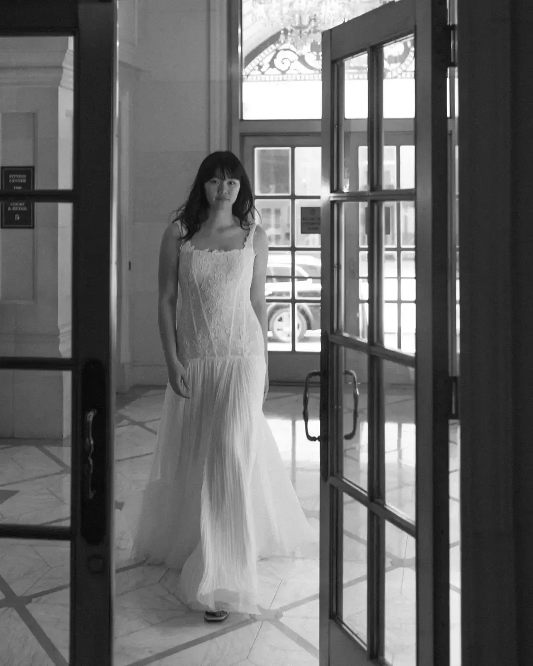 Woman in white dress, walking through doorway with glass panels, inside a building.