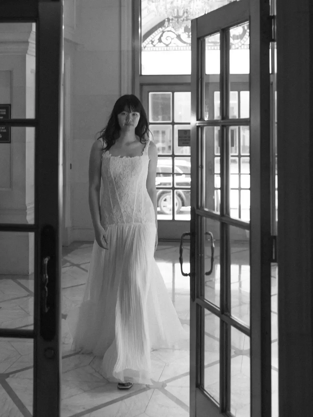 Woman in white dress, walking through doorway with glass panels, inside a building.