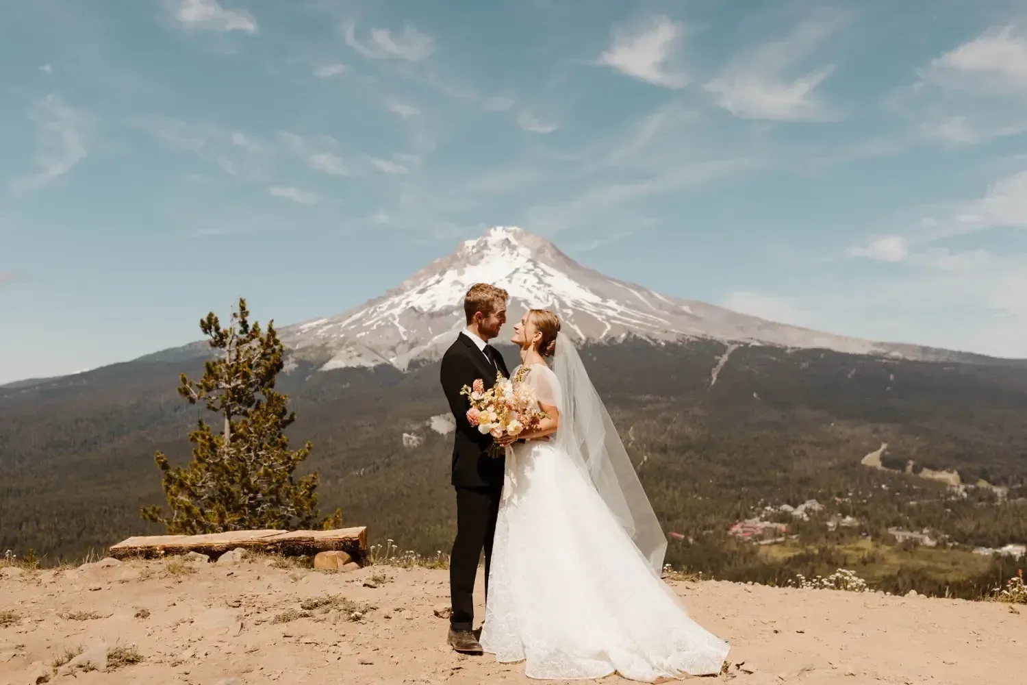 Couple embraces in wedding attire, overlooking a mountain. Bride in white dress, groom in black suit. Sunny sky.