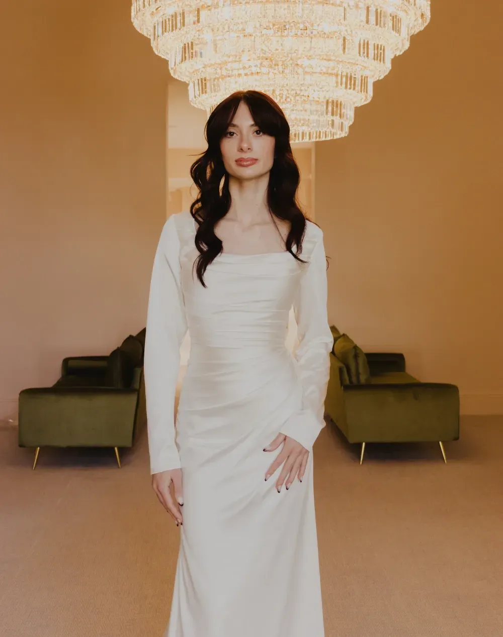 Woman in white dress, standing beneath a large chandelier, flanked by green velvet couches.