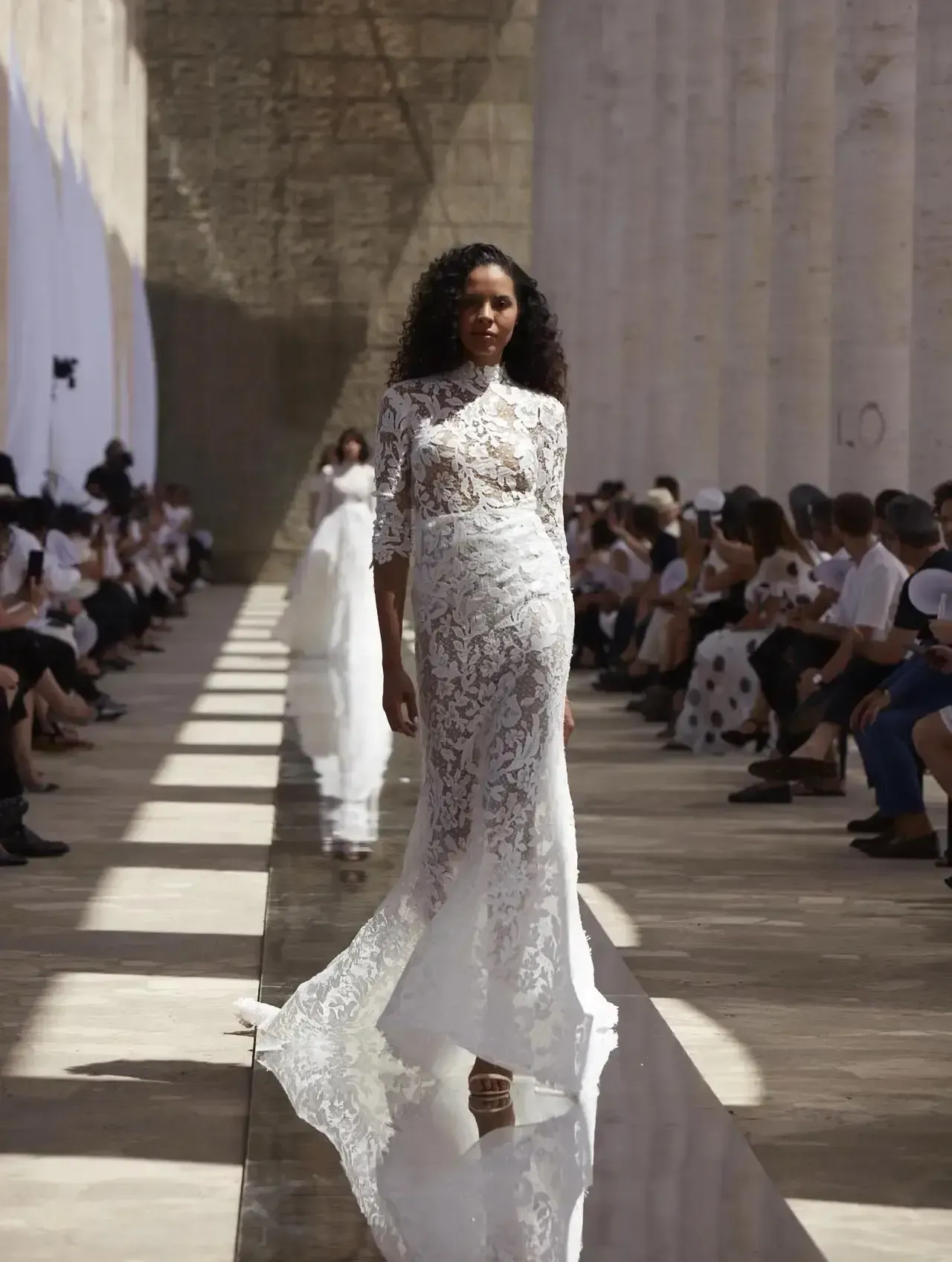 Woman in lace wedding dress walks runway between spectators.