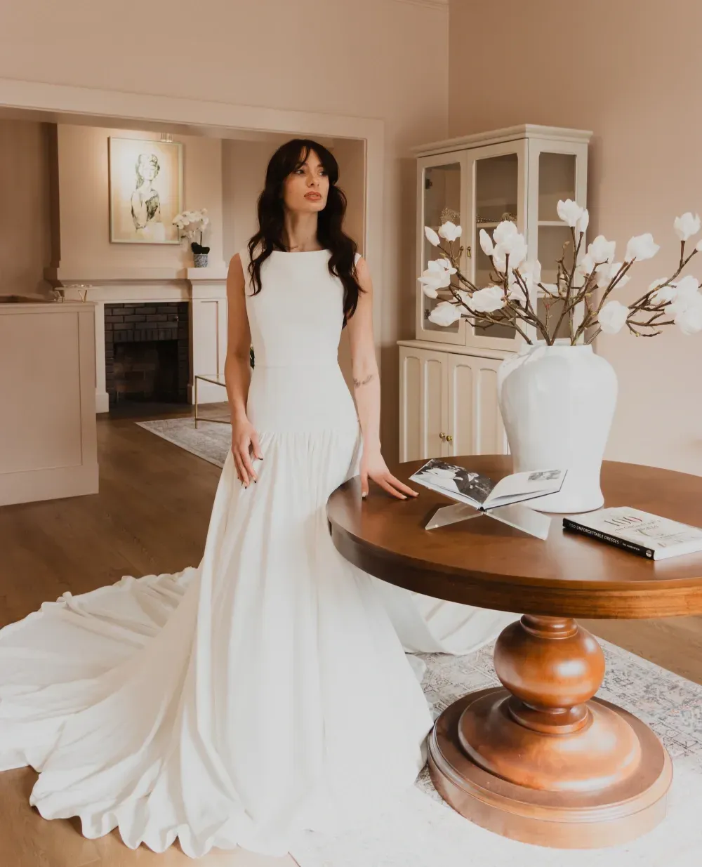 Woman in white wedding dress stands by a round table with flowers, indoors.