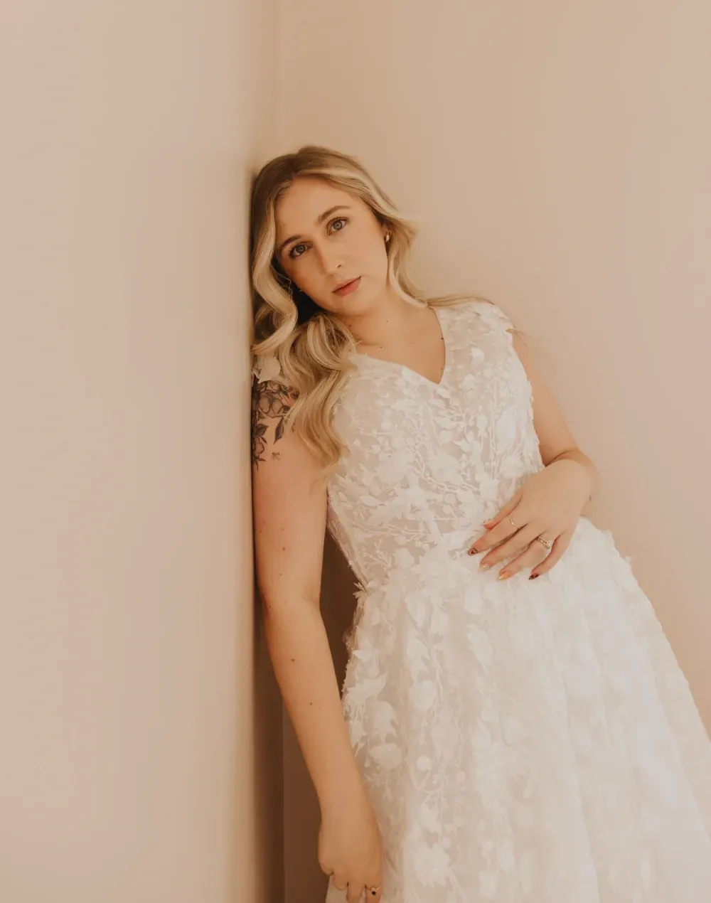 Blonde woman in white dress, leaning against pink wall, looking at the camera.