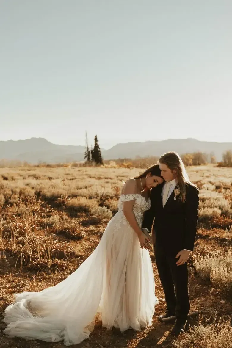 Bride and groom embrace in a field, mountains in background; sunlight, neutral tones.