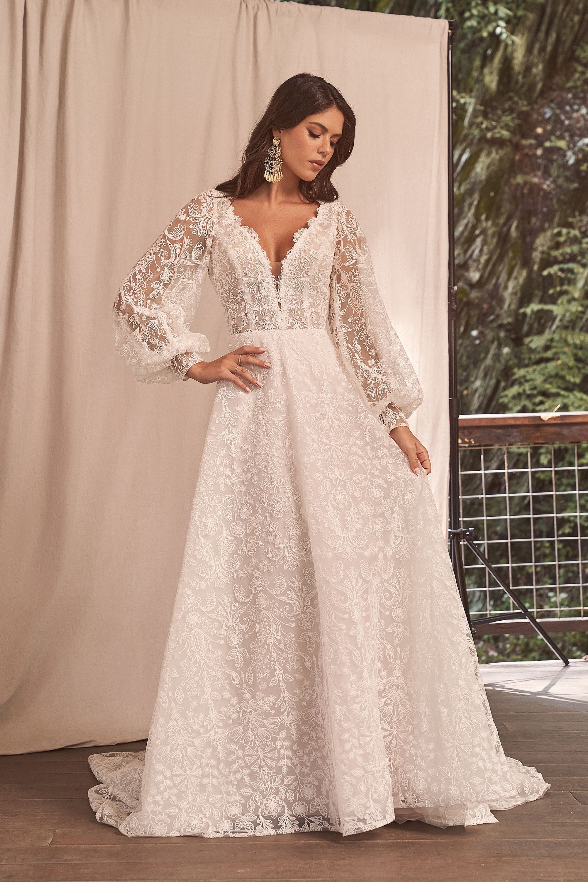 Woman in white, floral-embroidered wedding dress with long sleeves, posing outdoors.