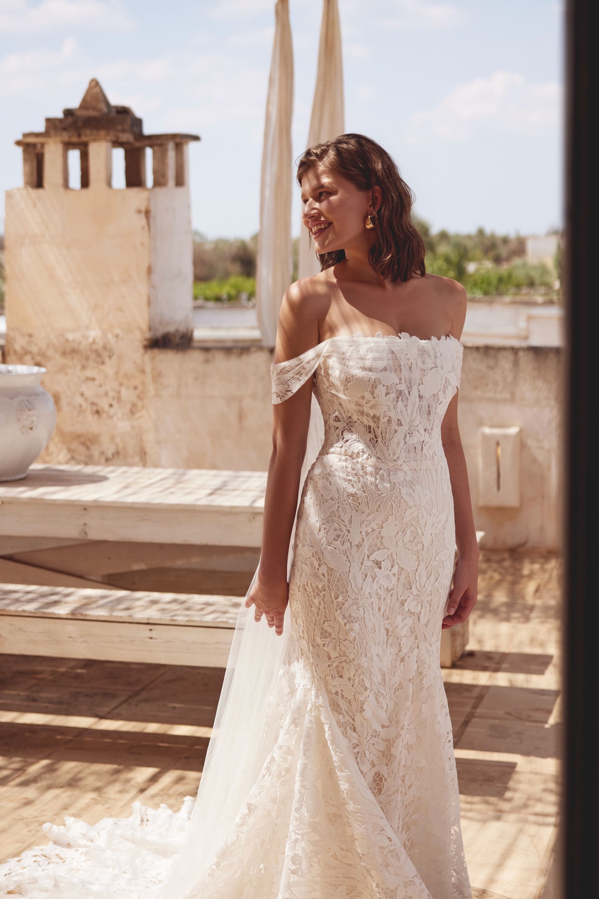 Woman in a lace off-the-shoulder wedding dress on a sunny terrace.