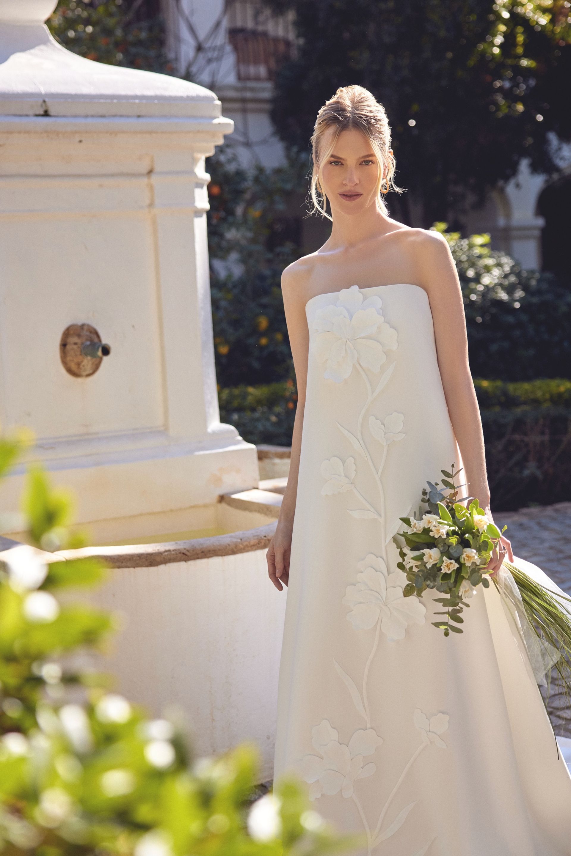 Woman in strapless white wedding dress holding bouquet, standing by a fountain in a garden.