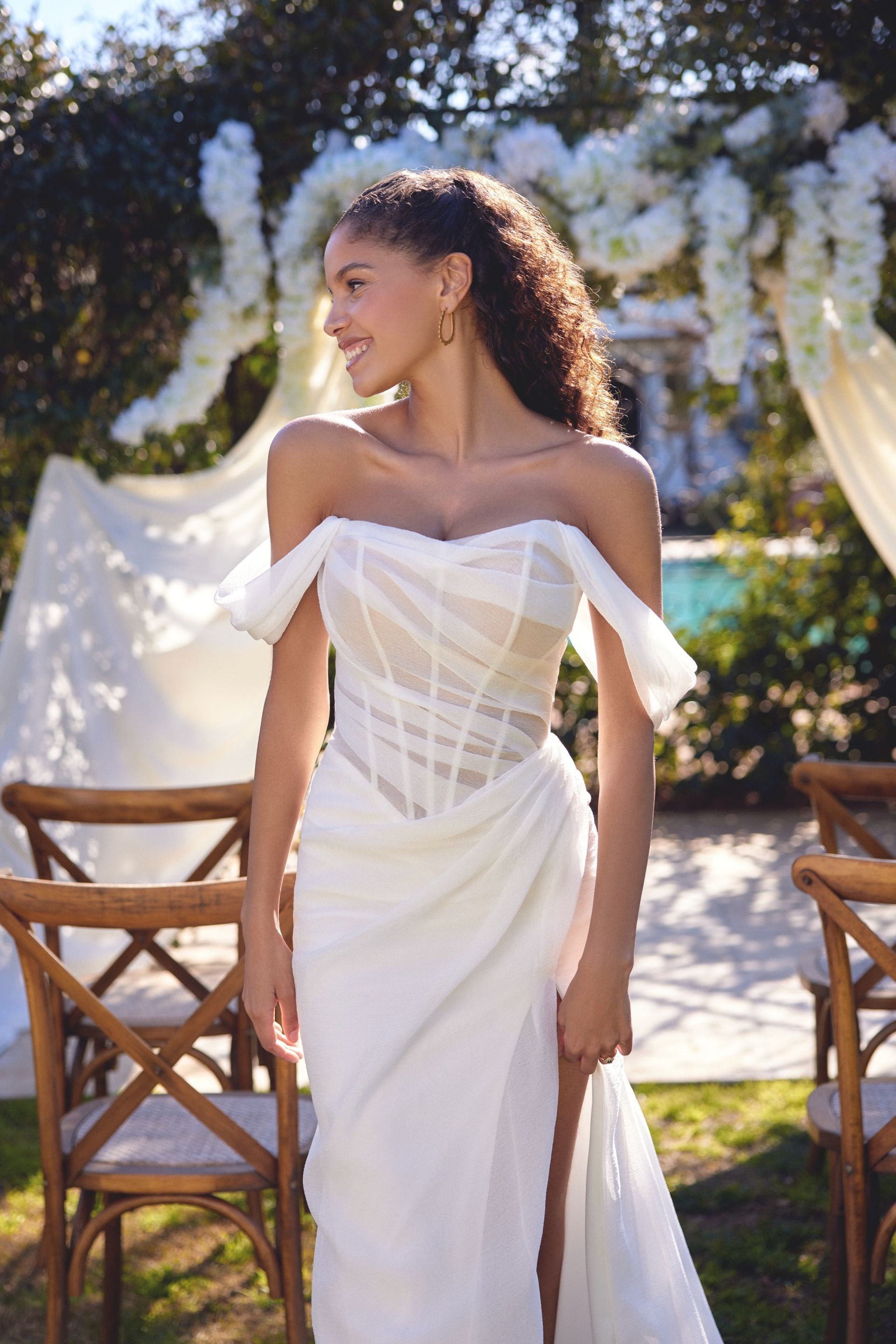 Woman in off-the-shoulder white wedding dress, smiling outdoors near ceremony setup.