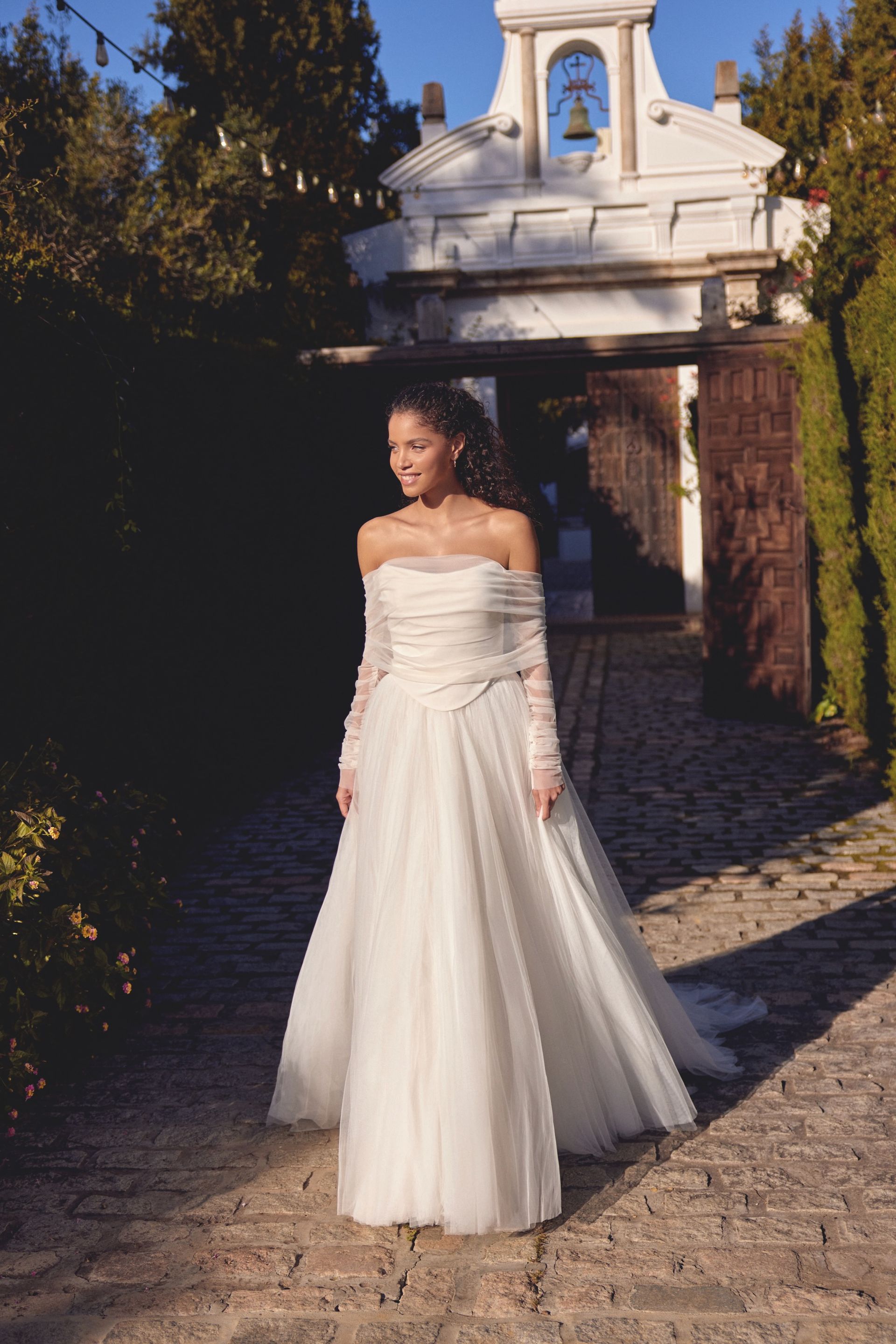 Woman in a wedding dress smiles, standing outdoors by a white ornate gate and a brick path.