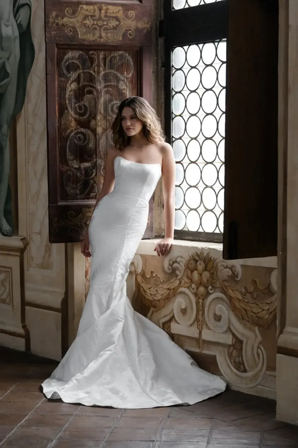 Woman in white strapless wedding gown leans against a decorated window.