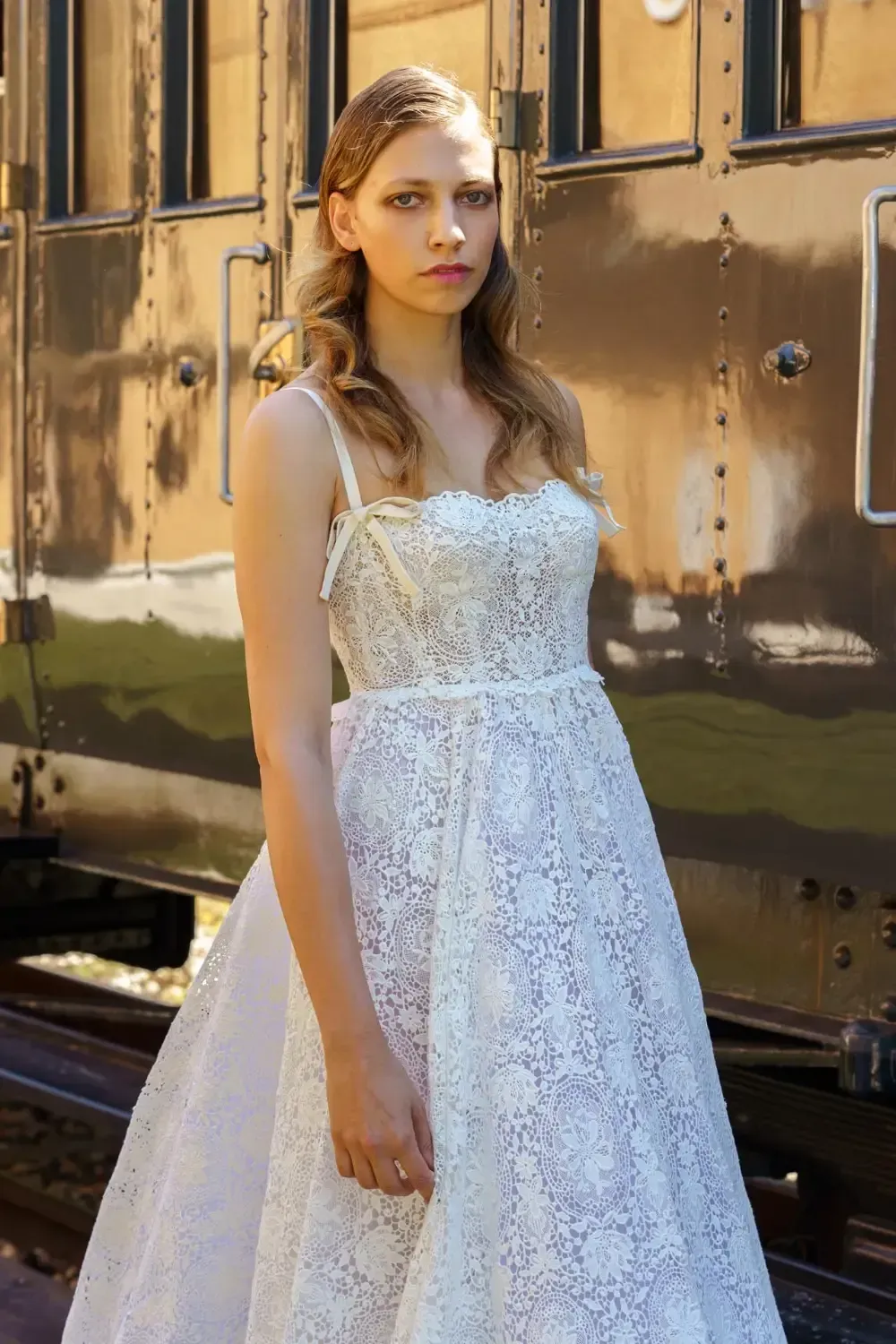 Woman in white lace dress poses by a train car.