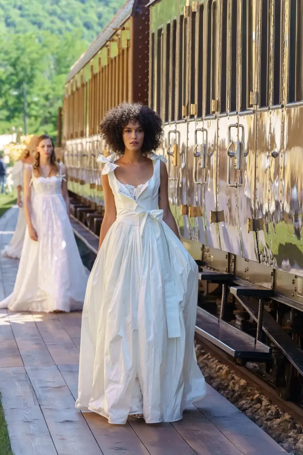 Woman in white dress walks on train platform, another woman in white dress in the background.