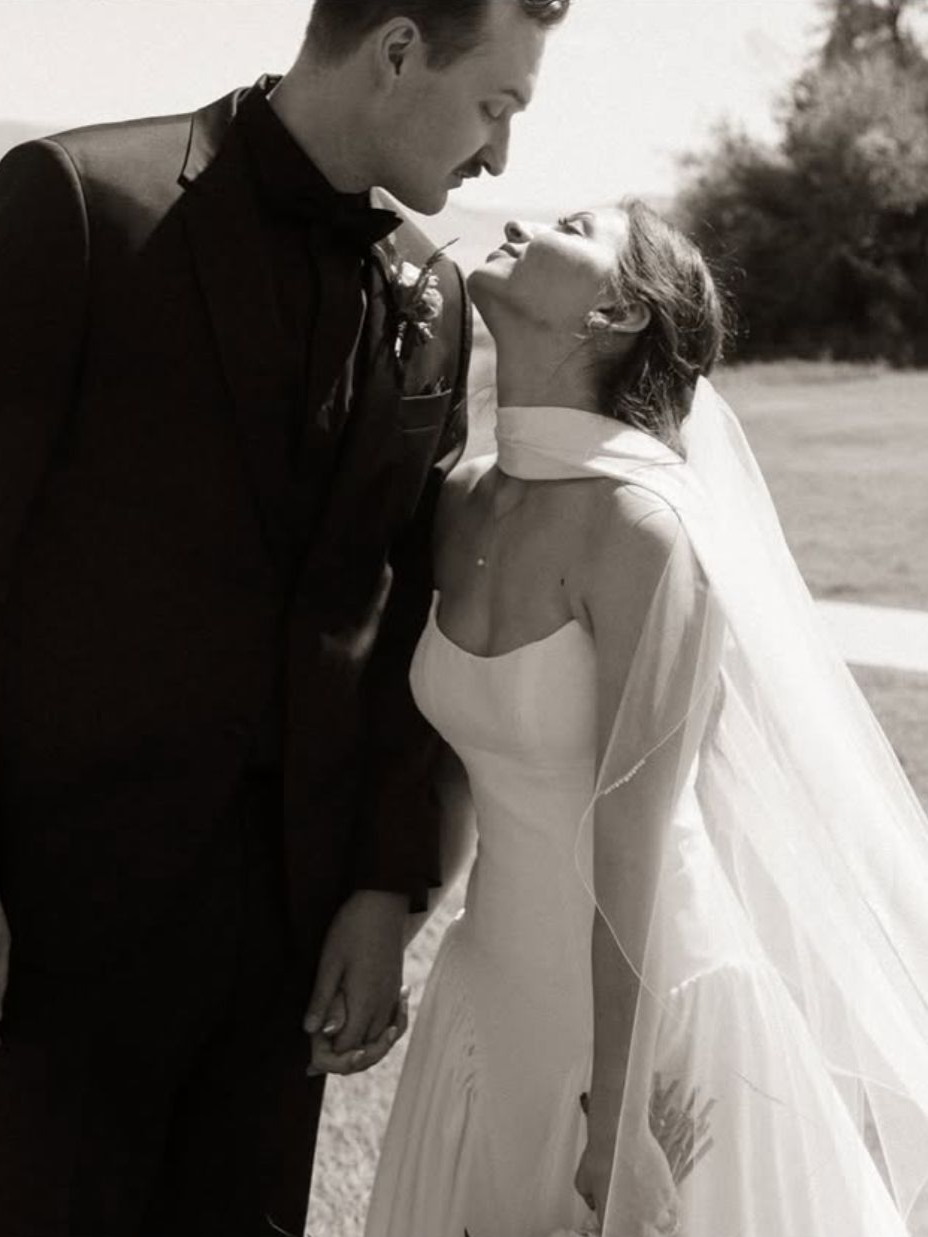 Newlyweds gazing at each other. Bride in white dress, veil. Groom in suit. Outdoors, sepia.
