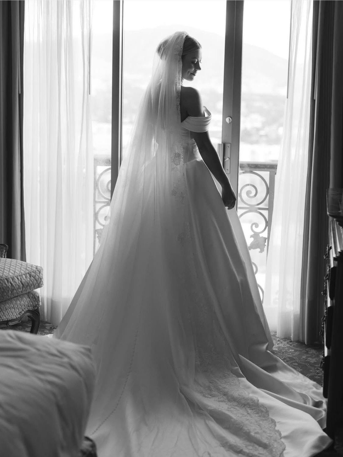 Bride in a wedding dress, standing by a window, with a long veil and train. Black and white.