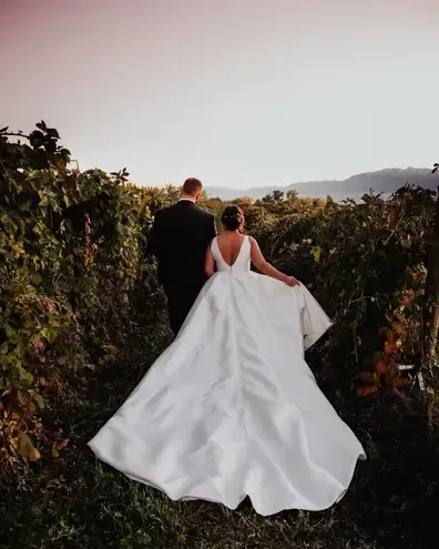 Groom carries bride in field, mountain backdrop. She wears white dress, he wears a suit, both smiling.