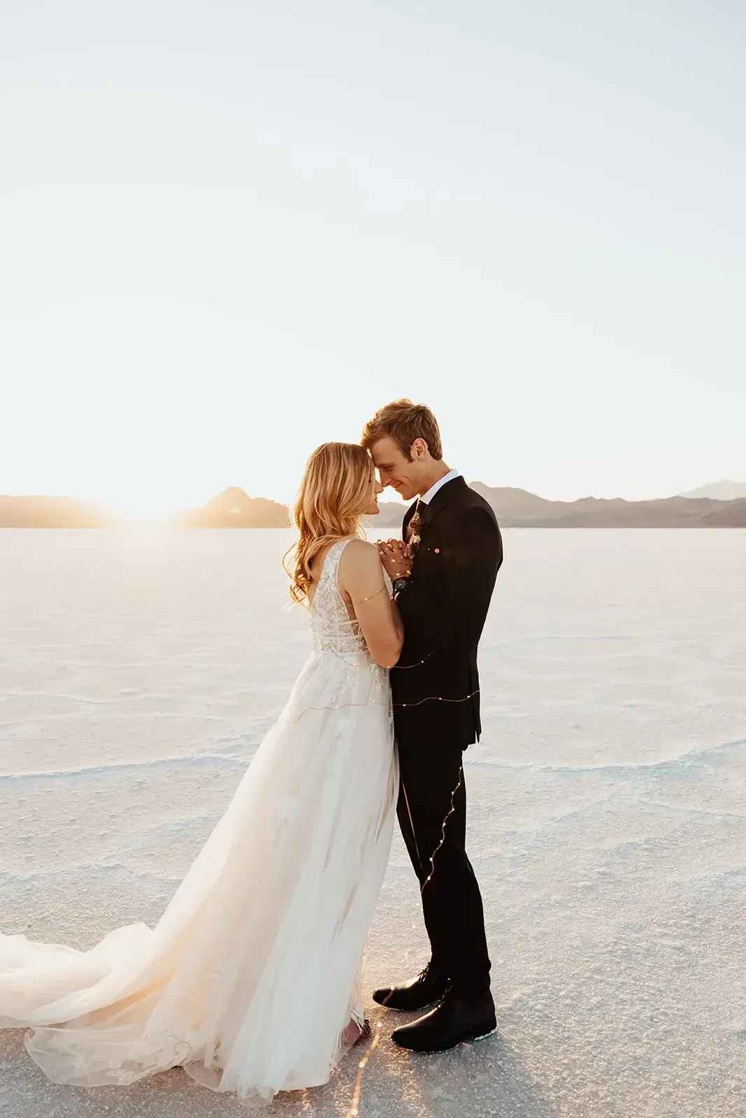 Couple embracing on a white salt flat at sunset; the bride in a white gown, the groom in a black suit.
