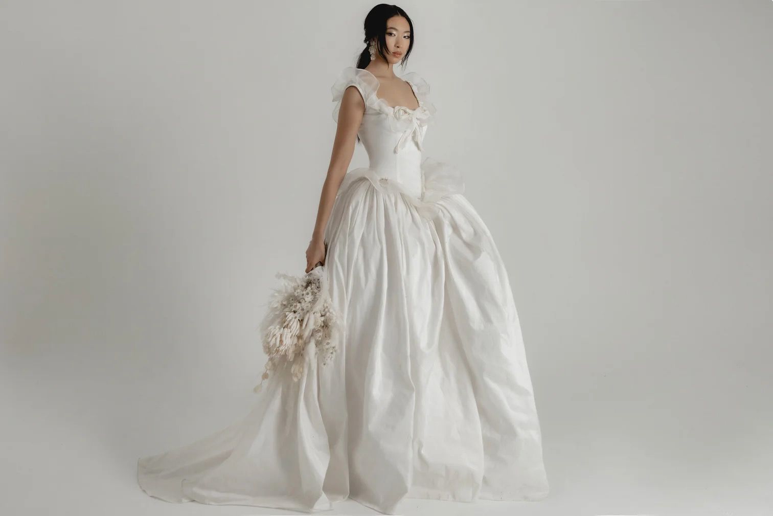Woman in a white wedding gown, holding bouquet, posing against a white background.