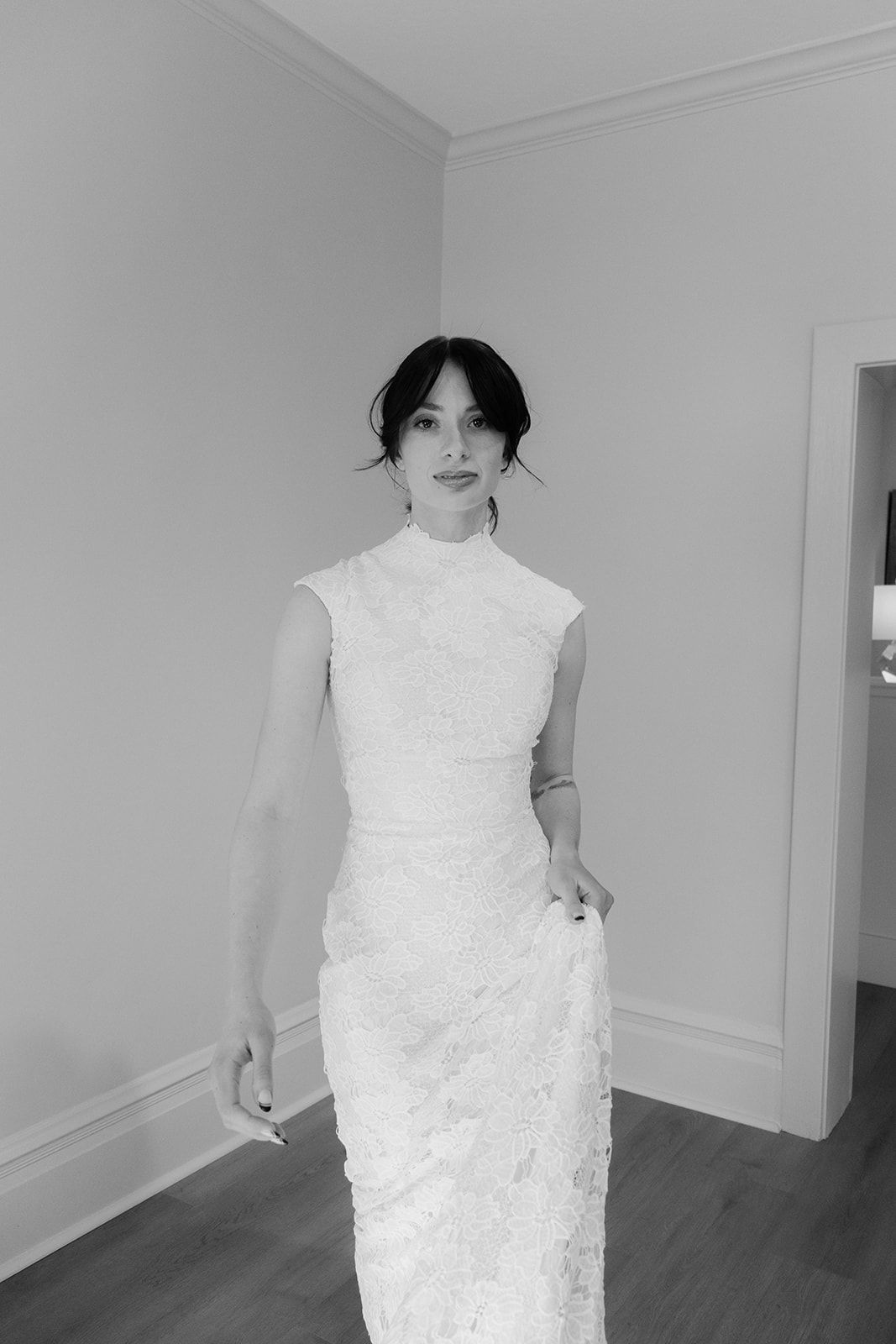 Woman in white lace dress, standing in a room. She has dark hair and a neutral expression.