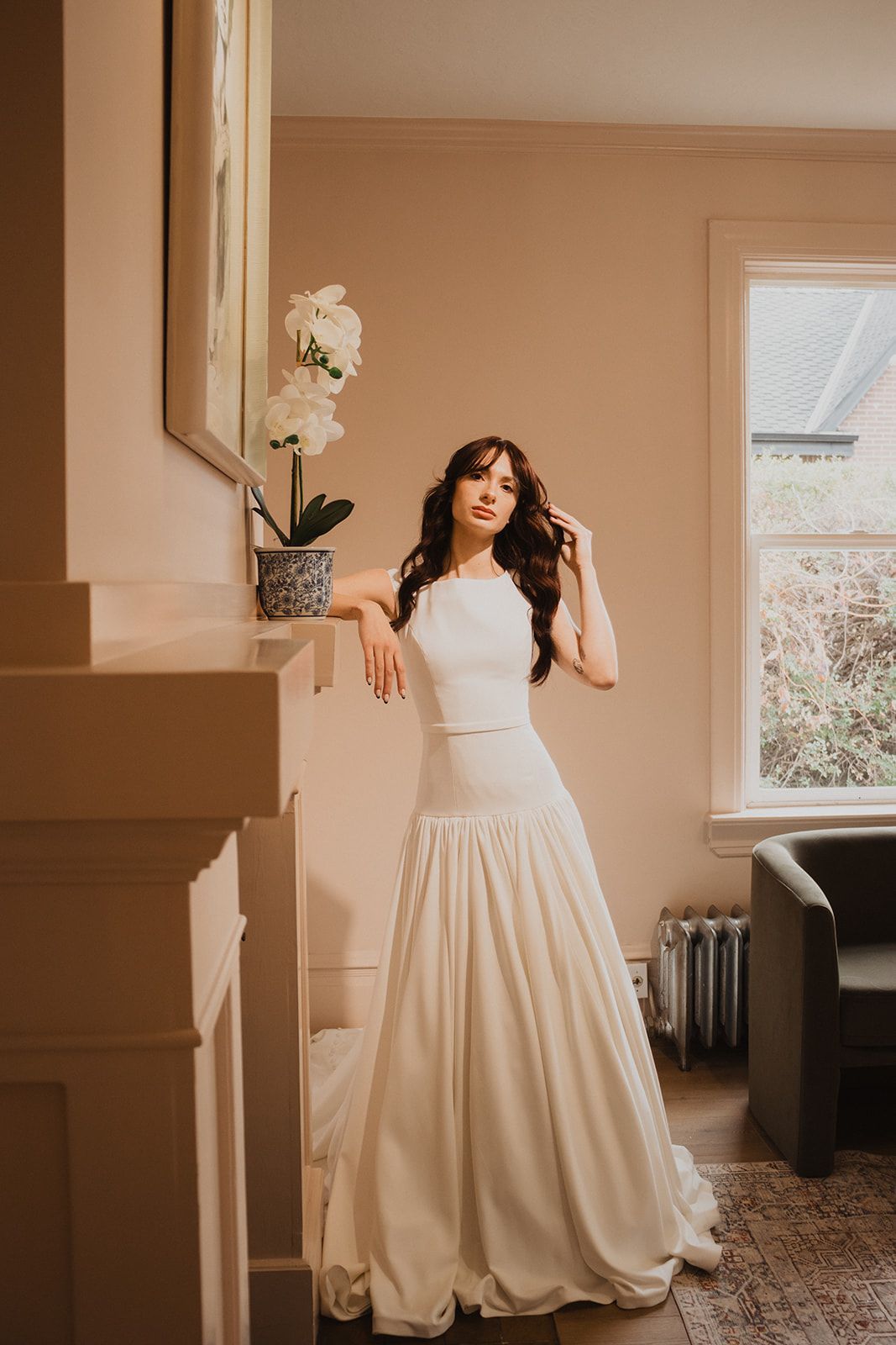 Woman in white gown leans on a mantel. Room with window and potted flowers.