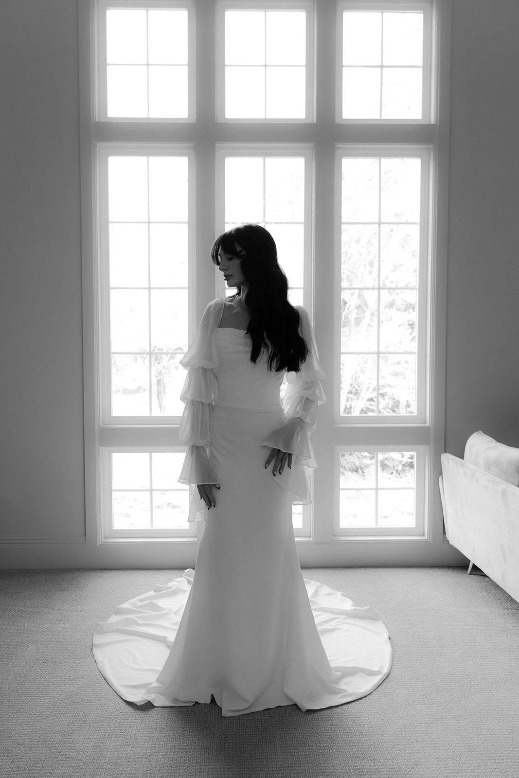 Bride in wedding dress, standing before a large window, with long train trailing on floor.