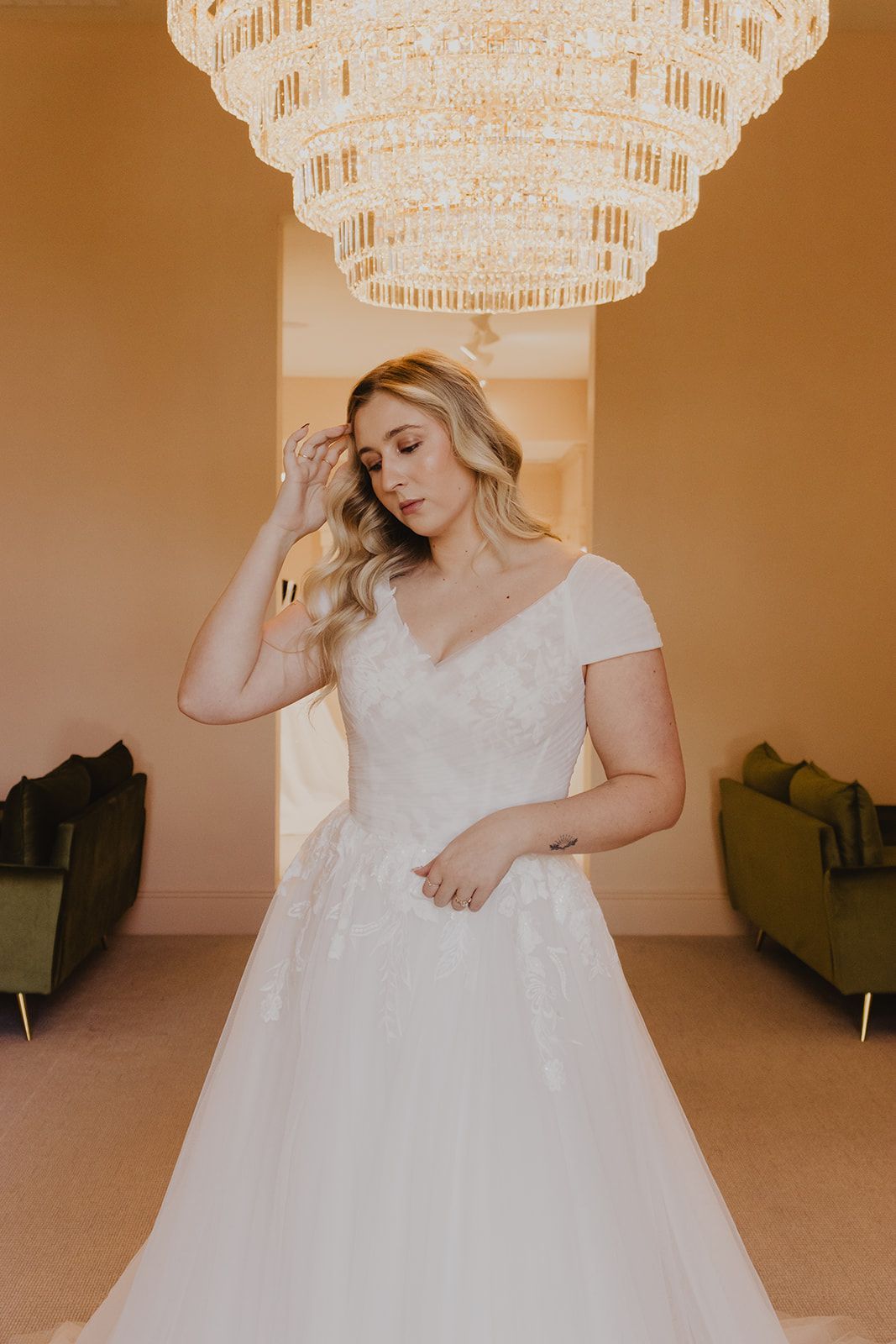 Woman in a white wedding dress stands under a chandelier, hand on her head, looking down.