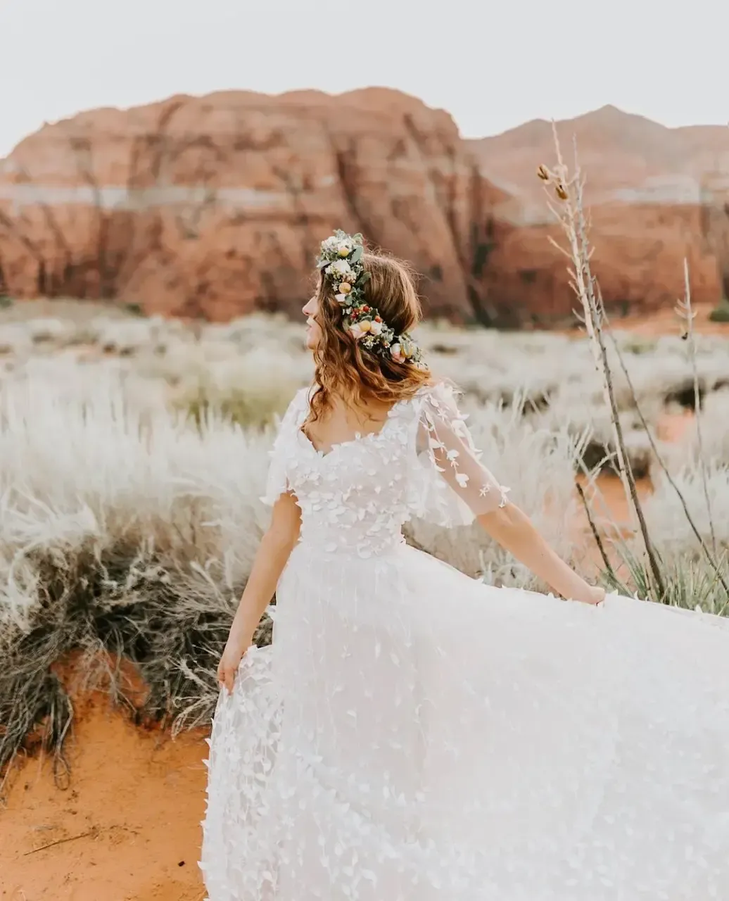 Woman in white floral wedding dress twirls outdoors, with red rock formations and dry grass.