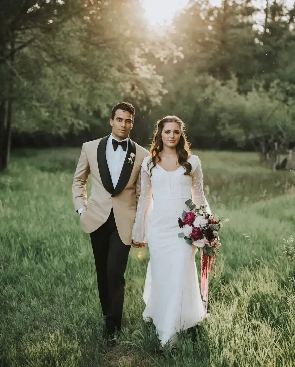 Couple walking in a grassy field; the man in a tan jacket, the woman in a white dress, holding hands, and carrying flowers.