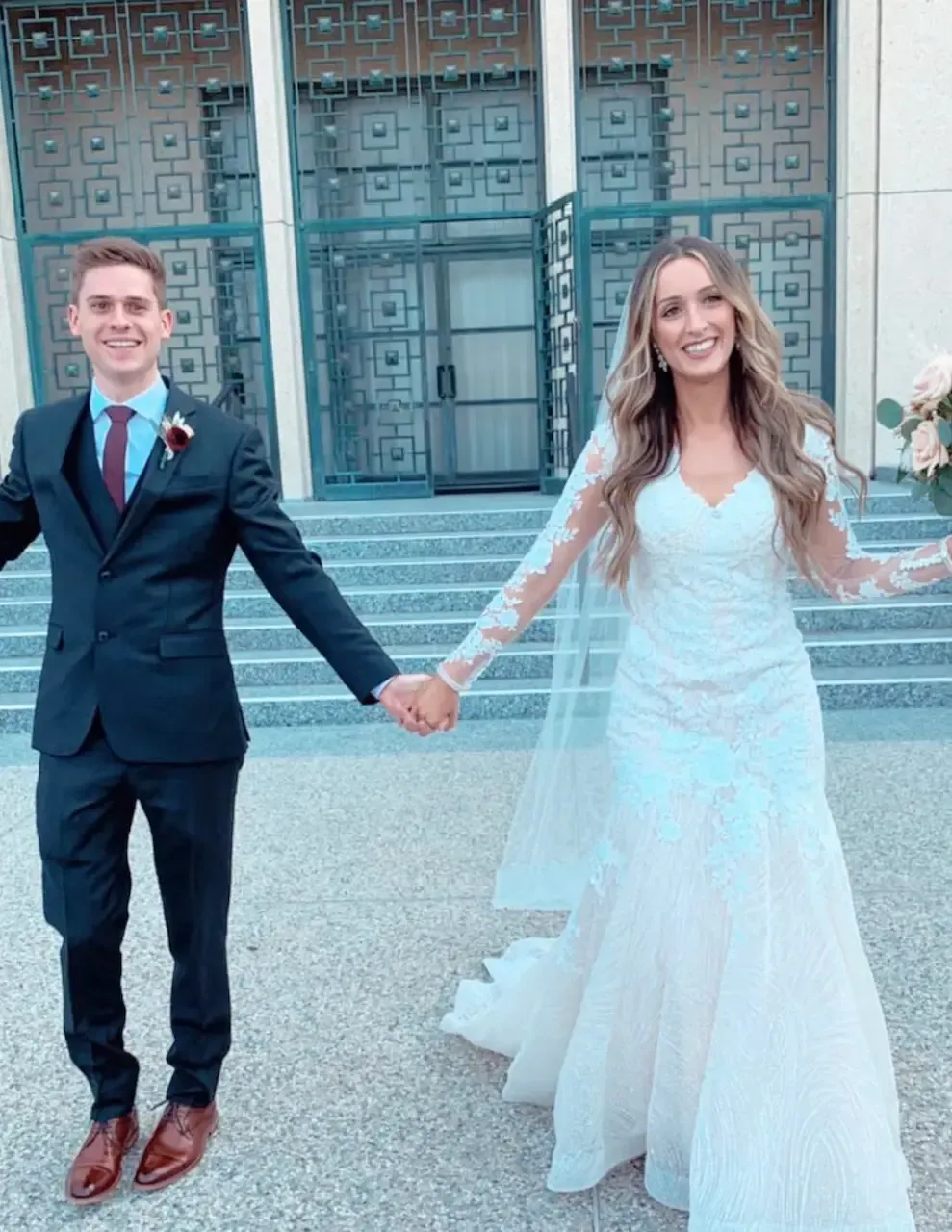 Newlyweds holding hands, smiling, in front of building with ornate doors. Woman in long-sleeve gown, man in suit.