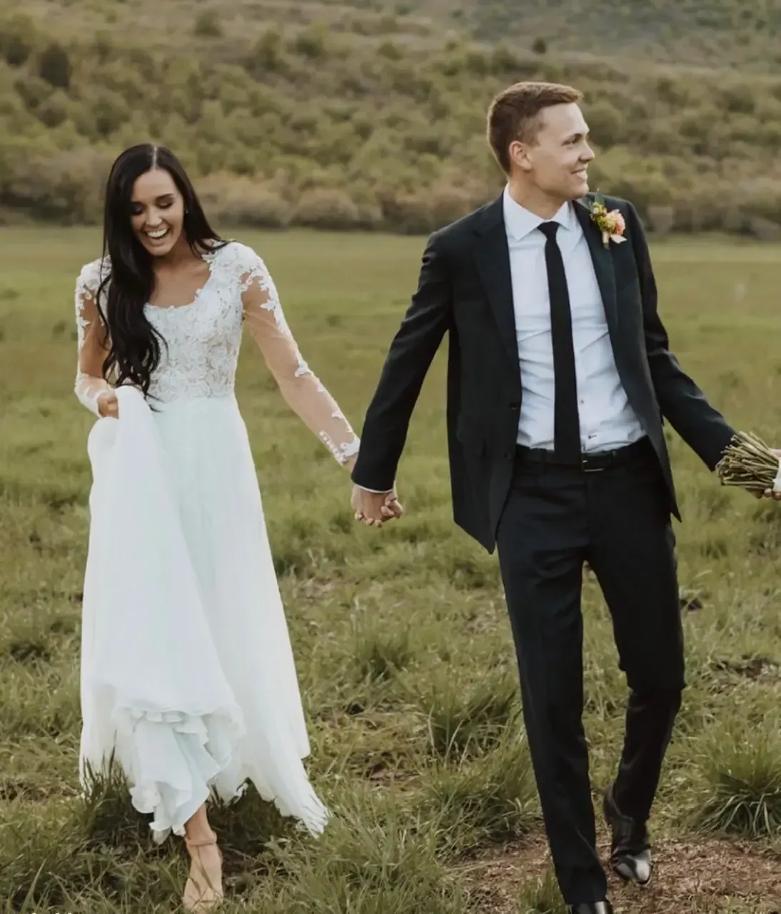 Bride and groom holding hands, smiling, walking in a grassy field. Bride in white dress, groom in black suit.