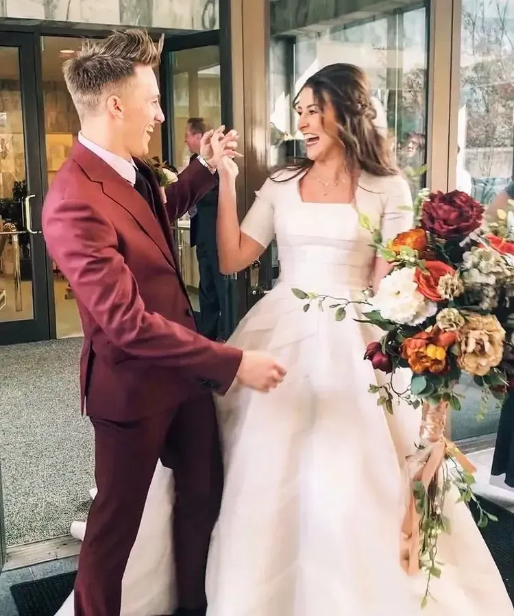 Bride and groom laugh, dancing outside a building after a wedding. Groom wears a maroon suit, bride a white dress.