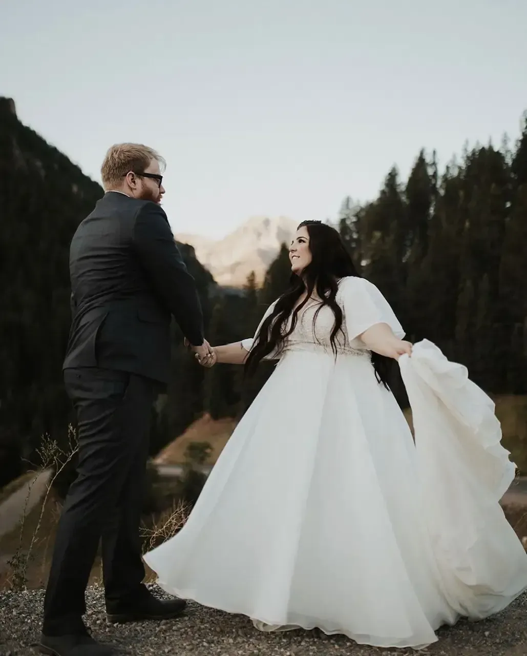 Couple holds hands, mountain backdrop. Woman in flowing white dress, man in suit.