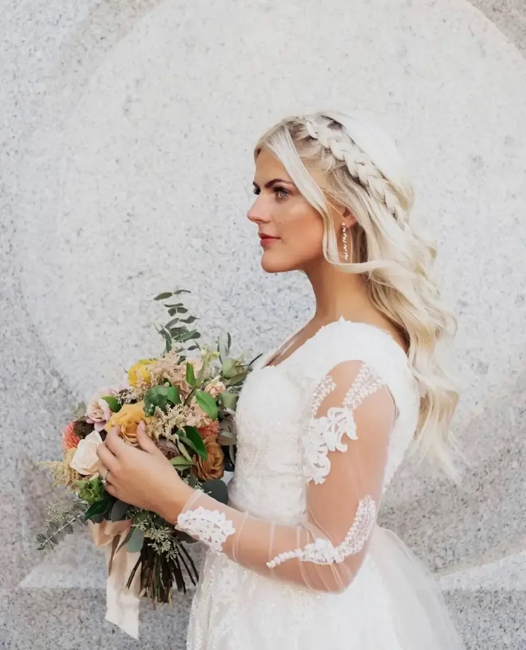 Blonde woman in wedding dress, holding flowers, looking to the side, against a stone wall.