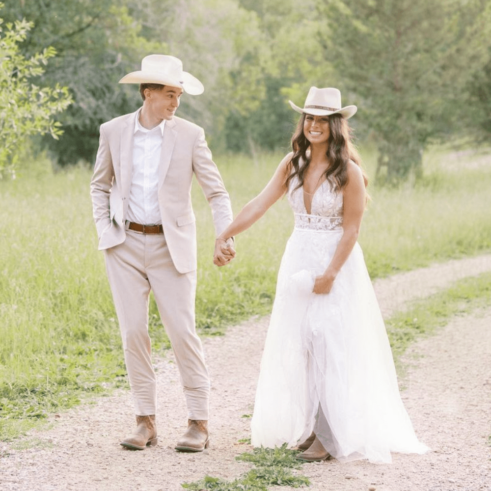 Couple in cowboy hats holding hands, walking on a dirt path in a field, smiling. Man in suit, woman in white dress.