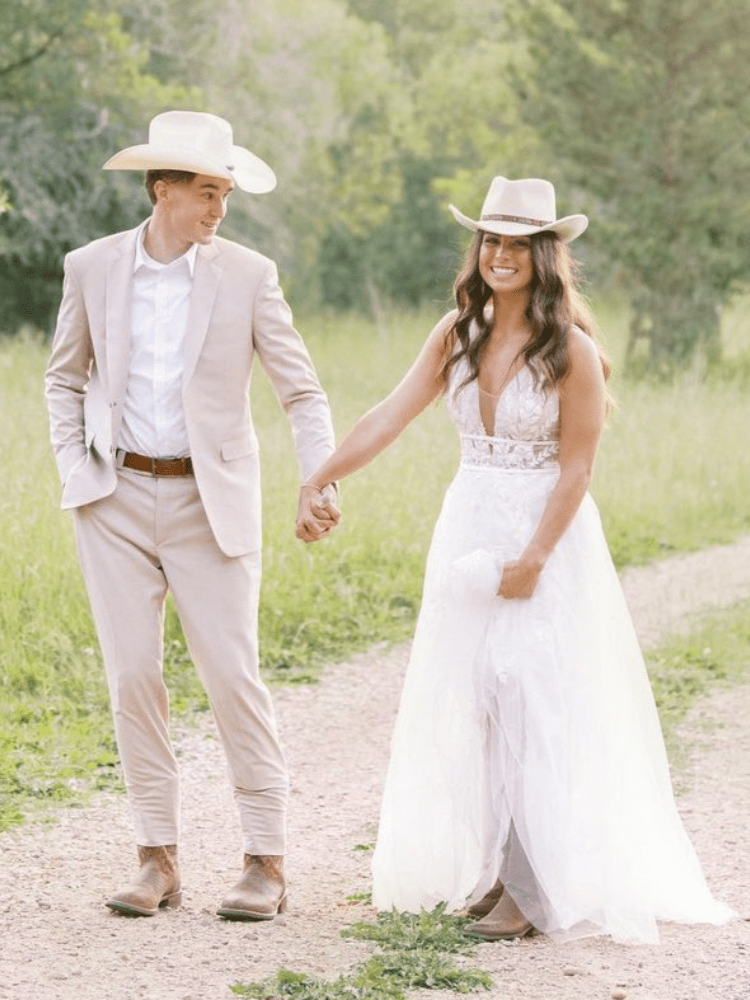 Couple in cowboy hats holding hands, walking on a dirt path in a field, smiling. Man in suit, woman in white dress.